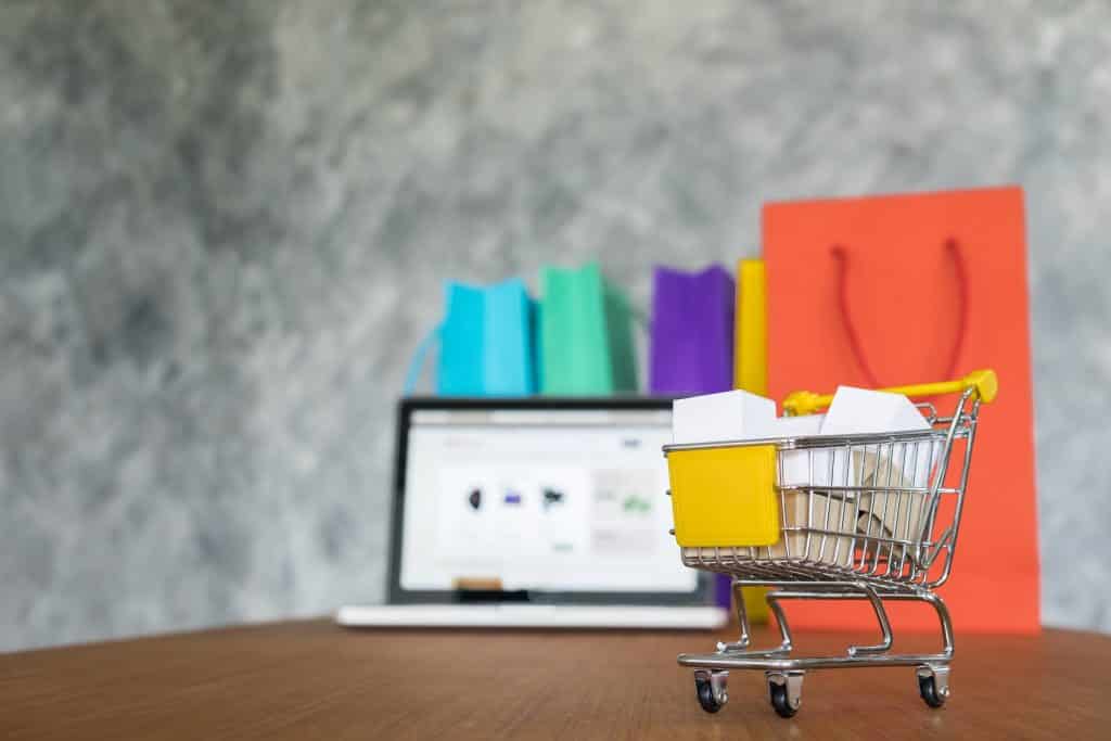 carrinho de compras com sacolas ao fundo e um computador. Marketplace é uma forma importante de vendas online