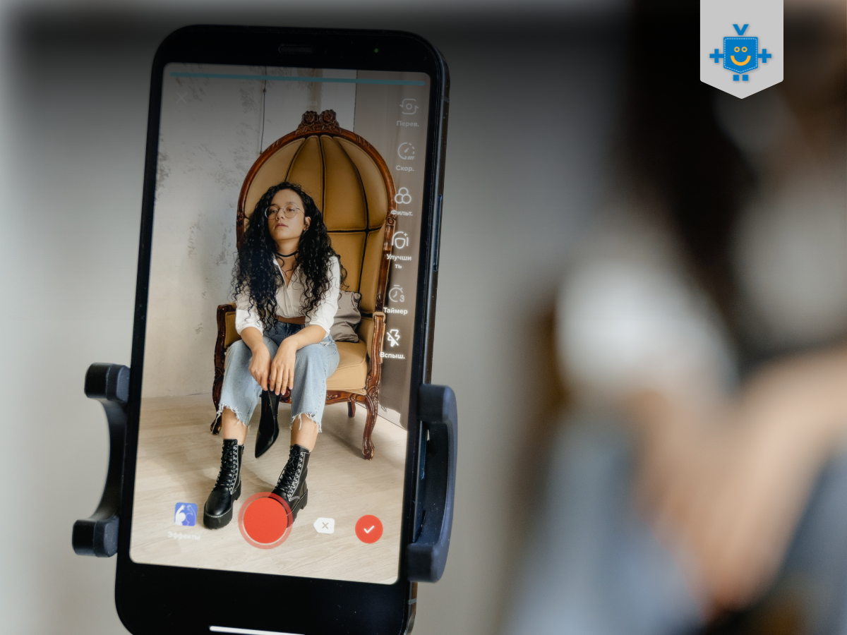 A imagem mostra uma criadora de conteúdo gravando um vídeo para a TikTok Shop. Ela aparece sentada em uma cadeira elegante, com o celular em um suporte, pronto para capturar o conteúdo. A interface do aplicativo TikTok está visível na tela, sugerindo a produção de um vídeo promocional ou de vendas.