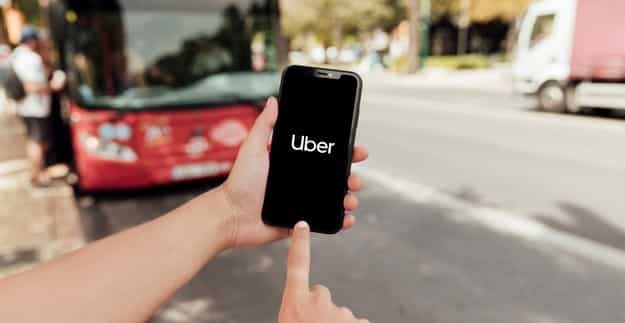 dedo apontando para um celular com o aplicativo uber aberto. ao fundo, uma rua com um ônibus vermelho